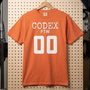 May include: An orange short-sleeve t-shirt featuring white screen-printed text that reads CODEX FTW above the large white numbers 00. The shirt has a crew neckline and is displayed on a wooden hanger against a light-colored pegboard background.