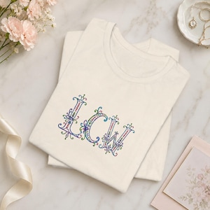 May include: A cream-colored short-sleeve t-shirt featuring the letters LCW embroidered in the center. The letters are decorated with intricate, colorful floral and vine patterns in shades of blue, green, pink, and purple with a filigree style.
