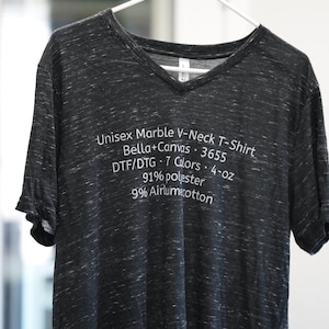 May include: A dark grey marble-patterned V-neck t-shirt with white text printed on the front that reads: Unisex Marble V-Neck T-Shirt, Bella+Canvas, 3655, DTF/DTG, 7 Colors, 4-oz, 91% polyester, 9% Airlume cotton. The shirt hangs on a white hanger.