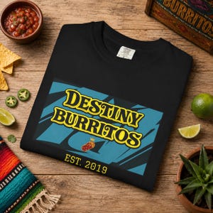 May include: A black t-shirt featuring a rectangular graphic with a blue background and black abstract shapes. The text reads "DESTINY BURRITOS" in yellow with a black outline, a small burrito icon with a crown, and "EST. 2019" in yellow text below.