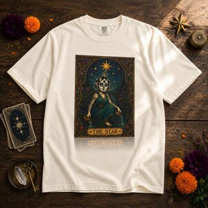 May include: Off-white t-shirt featuring a tarot card design. The card depicts a figure with face paint, a star, and the text "THE STAR". The design is framed with floral elements and a gold border. The shirt is displayed on a wooden surface.
