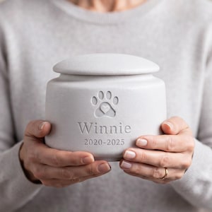 May include: A light grey cylindrical ceramic pet urn with a removable lid. The front features an engraved paw print with a small heart in the centre, the name "Winnie" below it, and the dates "2020-2025" at the bottom in a clean font.