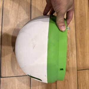 May include: A semi-spherical plastic bowl with a smooth white exterior and a bright green rim. The object features a small metal loop attached to the side of the green rim and a rectangular opening near the base of the white section.