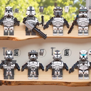 Könnte beinhalten: Eine Sammlung von acht weißen und grauen Actionfiguren, jede mit einzigartigen Helmdesigns und Zubehör. Die Figuren werden auf einem Holzregal präsentiert und zeigen verschiedene Waffen und Rüstungsteile. Die Figuren sind so gestaltet, dass sie Charakteren aus einem Science-Fiction-Franchise ähneln.