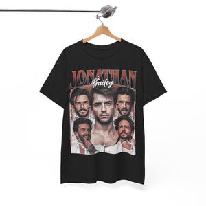 Pode incluir: Uma t-shirt preta de manga curta com uma impressão gráfica de Jonathan Bailey. O design inclui o nome JONATHAN em letras vermelhas grandes, o nome Bailey em escrita branca e cinco retratos fotográficos diferentes do ator.