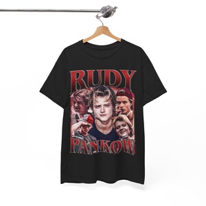 Op de afbeelding: Een zwart t-shirt met korte mouwen en een grafische print van acteur Rudy Pankow. Het ontwerp bevat meerdere fotografische portretten van de acteur in een collage, met de naam RUDY PANKOW in grote rode letters gedrukt.