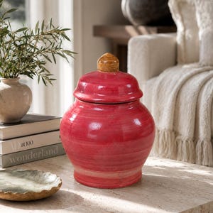 Puede incluir: Un jarrón de cerámica roja con tapa y pomo de madera. El jarrón tiene forma redondeada y superficie texturizada. Se encuentra sobre una mesa clara, sugiriendo un elemento decorativo para el hogar.