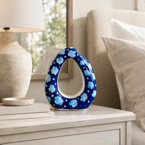 May include: A blue ceramic vase with a unique, open design. The vase is adorned with light blue floral patterns and white dots. It sits on a light-colored wooden nightstand, next to a lamp and a small dish. The vase is approximately 10 inches tall.