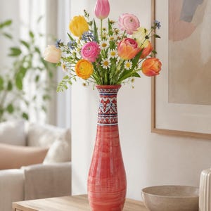 May include: A tall, red ceramic vase with a patterned neck holds a vibrant bouquet of colorful flowers. The arrangement includes tulips, ranunculus, and daisies. The vase sits on a wooden surface, with a neutral-toned painting and a bowl in the background.