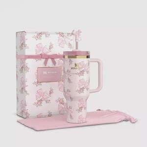 May include: A light pink Stanley tumbler with a floral and bow pattern, featuring a matching straw with a bow topper, a handle, and a gold-tone rim. It is presented with a rectangular gift box and a soft pink fabric pouch, both matching the design.