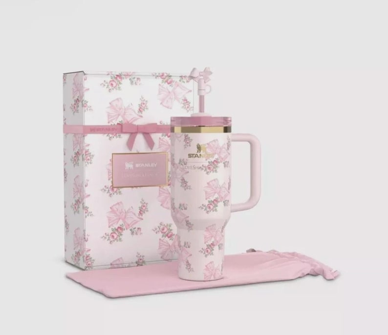 May include: A light pink Stanley tumbler with a floral and bow pattern, featuring a matching straw with a bow topper, a handle, and a gold-tone rim. It is presented with a rectangular gift box and a soft pink fabric pouch, both matching the design.