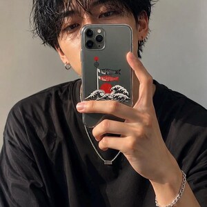 May include: A clear smartphone case featuring a graphic illustration of two koi fish streamers flying on a pole above stylized white ocean waves and a large red sun circle. The design incorporates black, white, and red colors on a transparent background.