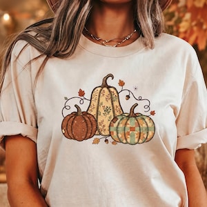 May include: A beige short-sleeve t-shirt featuring a graphic print of three pumpkins. The center pumpkin is tall with a leaf pattern, while the side pumpkins have textured brown and green patchwork designs, accented by small autumn leaves and acorns.