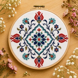 May include: A circular wooden embroidery hoop containing white fabric with a cross-stitch pattern. The design features a central dark blue star-shaped flower surrounded by symmetrical red tulip motifs, teal floral elements, and dark blue vines.