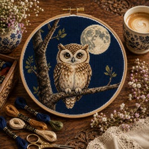 May include: A circular wooden embroidery hoop containing a cross-stitch design of a brown owl perched on a tree branch against a dark blue night sky with a full moon. The owl has large eyes and detailed plumage, rendered in shades of brown and tan.