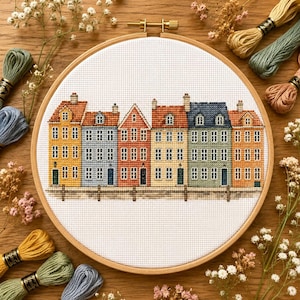 Puede incluir: Un bastidor de bordado circular de madera que contiene un patrón de punto de cruz de seis casas adosadas coloridas en amarillo, azul claro, naranja, crema, verde y bronceado. Las casas cuentan con múltiples ventanas y puertas oscuras.