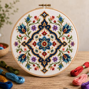Puede incluir: Un bastidor de bordado circular de madera que contiene tela blanca con un patrón de punto de cruz simétrico. El diseño presenta un motivo floral central azul y rojo rodeado de flores azules, rojas y moradas con tallos y hojas verdes.