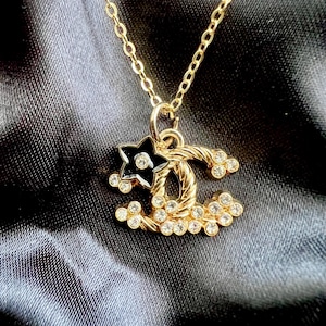 Puede incluir: Un collar de tono dorado con un colgante en forma de logotipo CC entrelazado. El colgante está adornado con numerosos cristales transparentes pequeños y un detalle en forma de estrella negra con un cristal central, en una cadena fina.