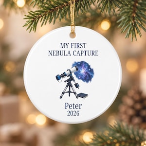 May include: A round white ceramic ornament featuring a graphic of a telescope pointed at a blue nebula. The text reads MY FIRST NEBULA CAPTURE, Peter, and 2026 in dark blue lettering. A gold string is attached to the top for hanging purposes.