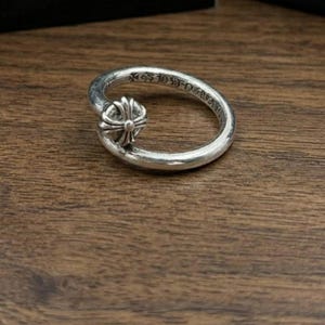 May include: A silver ring with a cross design on the front. The ring has a curved shape and is set against a wooden surface. The ring has engraved text around the band.