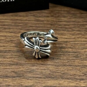 May include: A silver ring featuring a cross design and the engraved text "CHROME HEARTS" on the band. The ring has an intricate, decorative style and is displayed on a wooden surface.