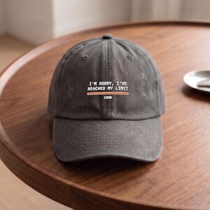 May include: A charcoal gray baseball cap featuring white text that reads I'M SORRY, I'VE REACHED MY LIMIT above an orange loading bar graphic with the text 100% underneath. The cap has a structured crown, a curved brim, and stitched eyelets.