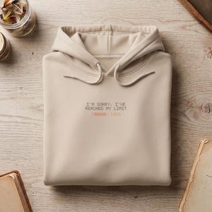 May include: A beige hooded sweatshirt featuring a black printed graphic that reads I'M SORRY, I'VE REACHED MY LIMIT above a loading bar icon with the text 100%. The garment includes a hood with matching drawstrings and a front kangaroo pocket.