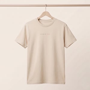 Könnte beinhalten: Ein beiges T-Shirt mit kurzen Ärmeln und Rundhalsausschnitt, das an einem Kleiderbügel hängt. In der Mitte der Brust befindet sich der kleine gestickte Text "I'M OUT OF FUCKS" in einer einfachen Schrift, wobei das letzte Wort "FUCKS" in zartem Rosa gestickt ist.