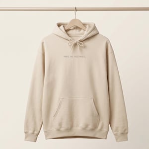 Op de afbeelding: Een beige hoodie met een kangoeroezak aan de voorzijde en lange mouwen. De tekst "MAKE NO MISTAKES." is in een klein, donker, gepixeld lettertype in het midden op de borst gedrukt. De hoodie wordt getoond op een kledinghanger.