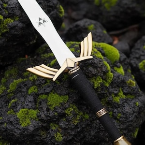 Può includere: Una spada color argento con una guardia dorata a forma di ali e un'impugnatura avvolta in nero. La lama presenta un simbolo Triforce triangolare nero vicino all'elsa. La spada è posizionata su una superficie rocciosa scura e strutturata.