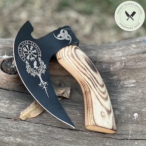 Könnte beinhalten: Ein schwarzes Metallbeil mit einer geschwungenen Klinge, die ein weiß graviertes Vegvisir-Symbol, einen Raben und ein Baummotiv zeigt. Der Griff besteht aus hellem Holz mit einer markanten dunklen Maserung. Ein Logo in der Ecke zeigt Trendy Blades Designs.