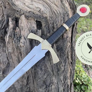 May include: A metal sword featuring a silver-toned blade, a gold-toned crossguard, and a black wrapped handle. The pommel is circular with a red gemstone inlay. A circular logo on the right displays the text TRENDY BLADES DESIGNS HANDMADE SHOP.