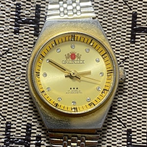 May include: A silver-tone analog wristwatch with a gold-colored dial featuring the text OREINTEX, three stars, and 17 JEWELS ANTIMAGNETIC. The dial has small clear stones as hour markers, gold hands, and a metallic link bracelet strap.
