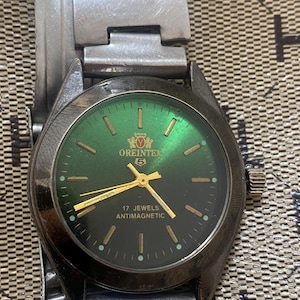 May include: A silver-toned wristwatch with a metallic green dial featuring gold-colored hour markers and hands. The watch face displays the text OREINTEX, a logo, the number 5, 17 JEWELS, and ANTIMAGNETIC. It includes a matching silver-toned metal link bracelet.