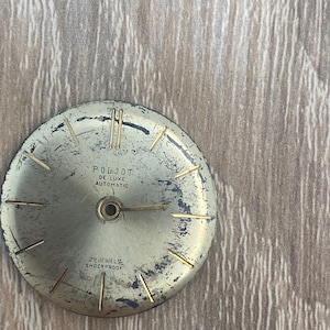 May include: A round, weathered gold-toned watch dial featuring thin gold hour markers and a single gold hand. The text reads Poljot De Luxe Automatic, 29 Jewels, Shockproof, and Made In. The surface shows significant wear, scratches, and dark patina.