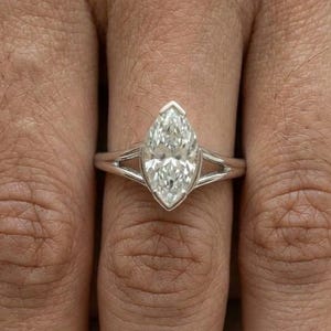 May include: A silver-toned ring featuring a marquise-cut clear gemstone set in a bezel setting. The band has a split-shank design that tapers towards the centre stone, creating a sleek and polished appearance on the finger.