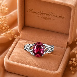 May include: A silver ring featuring a large, oval-cut, deep red gemstone set in a band with intricate, interwoven metalwork. The ring is displayed inside a tan velvet jewelry box with the text AmarJewelTreasure printed on the inner lid.