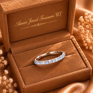 Op de afbeelding: Een zilverkleurige eternity ring met een doorlopende rij kleine, heldere en blauwachtig getinte stenen. De ring wordt gepresenteerd in een bruin fluwelen juwelendoosje met de tekst Amar Jewel Treasure US in goud op het deksel gedrukt.