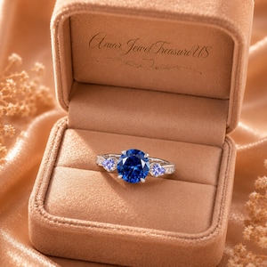 May include: A silver-toned ring featuring a large, round, deep blue center stone flanked by two smaller, pear-shaped, light blue stones, presented in a tan velvet box with the text Amar Jewel Treasure US printed on the inner lid.