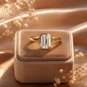 Puede incluir: Un anillo de oro con una gran piedra central rectangular de corte esmeralda en un engaste de bisel. La banda tiene un diseño de vástago dividido que se estrecha hacia la piedra. El texto "Amar Jewel Treasure" es visible en la esquina inferior derecha.