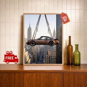 May include: A framed wall art print depicting a bronze-colored sports car suspended by chains against a New York City skyline featuring the Empire State Building and the Chrysler Building. The artwork is displayed next to two empty glass bottles.