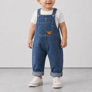 May include: A pair of blue denim overalls featuring a small brown bear patch on the chest pocket, worn over a plain white short-sleeved t-shirt. The overalls have metal buttons at the shoulders and are paired with white canvas sneakers.