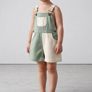 May include: Short overalls featuring a color-blocked design with one side in sage green and the other in off-white. The garment includes a front chest pocket, two side pockets, and adjustable shoulder straps with metallic buckles and buttons.