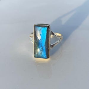 May include: A silver ring featuring a rectangular labradorite gemstone with a vibrant blue iridescent flash. The stone is set in a smooth metal bezel, and the band consists of two thin silver wires that meet at the sides of the setting.