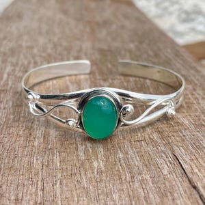 Peut inclure: Un bracelet manchette en argent orné d'une pierre précieuse verte de forme ovale au centre. La bande métallique présente une finition polie avec des détails en fil torsadé décoratifs de chaque côté du sertissage, créant un design ouvert.