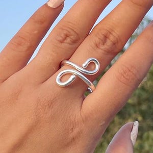 May include: A silver-toned ring featuring a continuous wire design that forms two symmetrical, rounded loops that meet in the center. The metal has a smooth, polished finish and an open-ended structure that wraps around the finger.