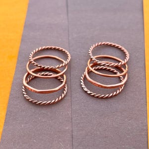 May include: Two sets of three copper-colored rings arranged in two stacks. Each stack contains two thin twisted rope-textured bands and one smooth, slightly hammered band. The rings have a metallic finish and a circular shape.