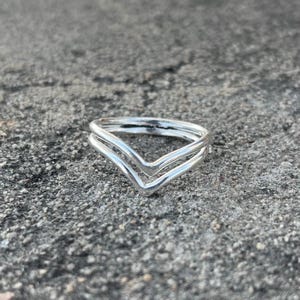 Può includere: Un anello in argento lucido caratterizzato da un design a doppia fascia a chevron o a forma di V. L'anello è composto da due sottili fasce metalliche lisce che si incontrano in un punto centrale, creando una silhouette angolare simmetrica.