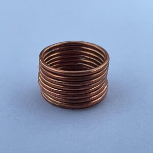 May include: A stack of ten thin, circular copper-colored rings. Each ring features a smooth, plain band with a metallic finish. The rings are stacked vertically, creating a cylindrical shape with a consistent diameter and uniform appearance.