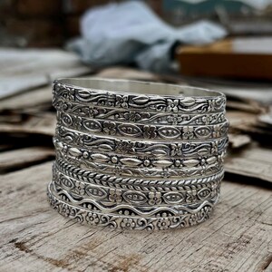 Peut inclure: Une pile de sept bracelets rigides en métal argenté présentant des motifs gaufrés complexes. Les designs incluent des motifs floraux répétitifs, des volutes délicates, des textures tressées et des formes géométriques avec une finition métallique polie.
